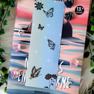 Puede incluir: Un marcapáginas azul claro con ilustraciones de mariposas y flores negras. El marcapáginas está colocado encima de un libro con una portada rosa y azul. Adornos decorativos en forma de rosa y luna creciente están dispersos.