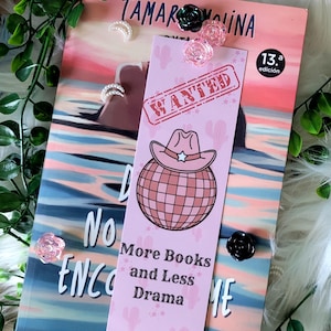 Puede incluir: Un marcapáginas rosa con un sombrero de vaquero sobre una bola de discoteca, con la palabra "WANTED" estampada y el texto "More Books and Less Drama". El marcapáginas está colocado sobre un libro con una portada colorida.