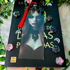 Puede incluir: Un libro negro con un marcapáginas que presenta el retrato de una mujer y una borla roja. El marcapáginas está colocado en un libro con el título "SARAH J. MAAS" visible. Alfileres decorativos iridiscentes en forma de rosa y alfileres en forma de media luna rodean el libro.