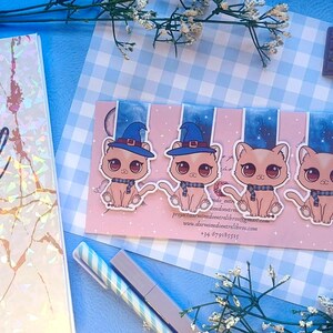 Puede incluir: Un conjunto de cuatro marcadores con temática de gatos. Cada marcador presenta un gato de dibujos animados con un sombrero de bruja y una bufanda. El fondo es una tarjeta rosa con un diseño de cielo estrellado azul.