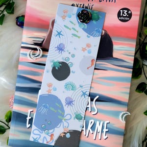 Puede incluir: Un marcapáginas decorativo con temática oceánica que presenta ilustraciones de criaturas marinas como medusas, caballitos de mar y estrellas de mar. El marcapáginas está adornado con un adorno negro en forma de rosa. El marcapáginas está sobre un libro.