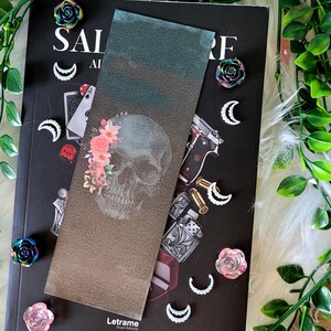 Puede incluir: Un marcapáginas con un diseño de calavera y flores, colocado sobre un libro negro. El marcapáginas tiene un degradado marrón y azul. Los elementos decorativos incluyen medias lunas, rosas e imágenes de naipes.