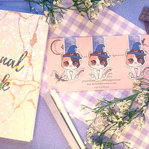 Puede incluir: Un diario con un diseño de mármol holográfico y las palabras "Journal book" en escritura negra. Tres pegatinas de gatos con sombreros de bruja están en una tarjeta rosa. El fondo incluye un patrón de cuadros morado y flores blancas.