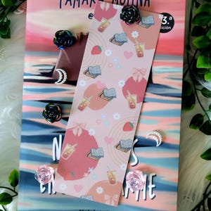 Puede incluir: Un marcapáginas rosa con un diseño de libros, té de burbujas, fresas y lazos, colocado sobre un libro. El libro tiene una portada colorida con texto. Alfileres decorativos en forma de rosa y luna están dispersos.