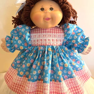 May include: A vintage doll with a round face, brown yarn hair, and blue eyes. The doll wears a blue floral dress with pink gingham trim and white lace accents. A white ribbon is tied in the hair.