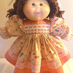 May include: A doll with brown yarn hair tied with white ribbons, featuring a round face, rosy cheeks, and a smiling mouth. The doll wears a peach-colored dress with a floral pattern, white lace trim at the bodice, and a polka-dot hemline.