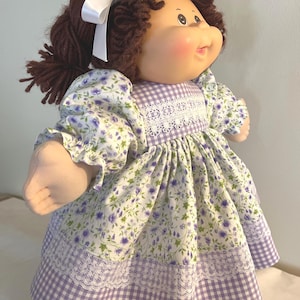 May include: A vintage doll with brown yarn hair, a white bow, and a rosy complexion. The doll wears a floral print dress with purple flowers, white lace, and a purple and white gingham hem. The doll's arms and hands are visible.