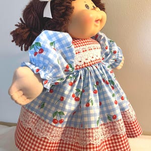 May include: A vintage doll with brown yarn hair and a white bow. The doll wears a blue and white gingham dress with red cherry accents and a red and white gingham skirt. The dress has white lace trim.