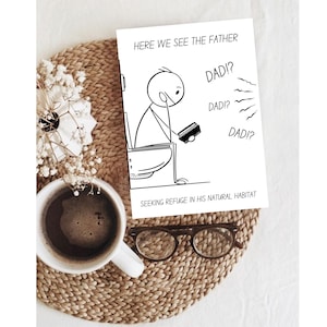 The humorous printable card featuring a stick figure father on a toilet is presented on a light brown woven placemat. A white mug of coffee and a small white floral arrangement are placed to the left, and a pair of reading glasses lies below the card. This warm, inviting setting suggests a relaxed environment, perfect for enjoying a thoughtful or humorous moment with the card. The overall composition is cozy and appealing, emphasizing the card's theme.