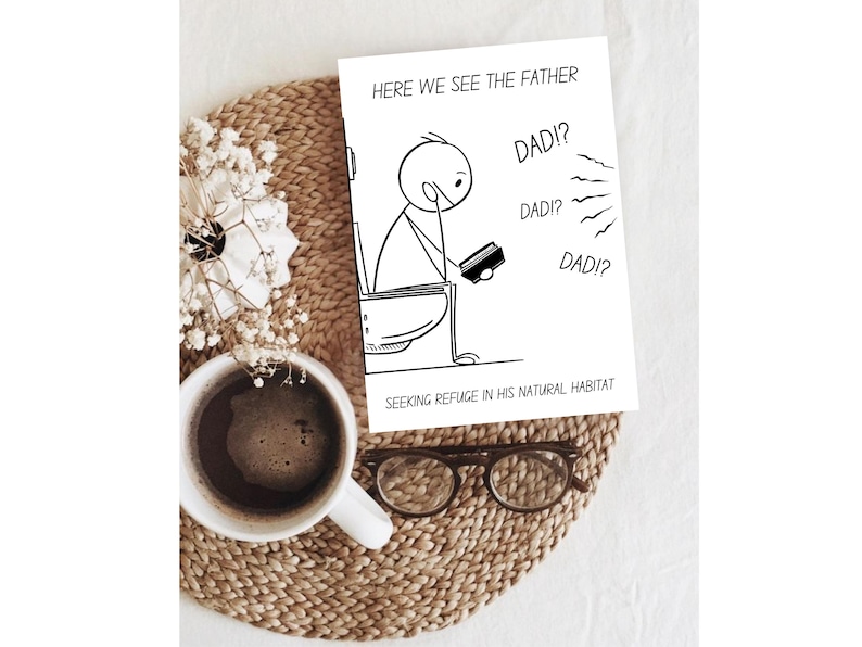 The humorous printable card featuring a stick figure father on a toilet is presented on a light brown woven placemat. A white mug of coffee and a small white floral arrangement are placed to the left, and a pair of reading glasses lies below the card. This warm, inviting setting suggests a relaxed environment, perfect for enjoying a thoughtful or humorous moment with the card. The overall composition is cozy and appealing, emphasizing the card's theme.