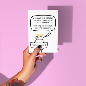 Peut inclure: Une carte de v&oelig;ux blanche avec le texte "WE HAVE THE PERFECT MOTHER-DAUGHTER RELATIONSHIP. YOU ARE MY MOTHER AND I'M PERFECT." Un personnage de dessin anim&eacute; avec un halo et une fleur tient un panneau "YOU'RE WELCOME."