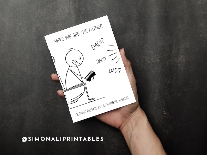 A hand holds the humorous printable card, which features a stick figure father on a toilet, against a dark gray background. The card's front displays the illustration and text "HERE WE SEE THE FATHER SEEKING REFUGE IN HIS NATURAL HABITAT" with "DAD!?" speech bubbles. The direct view emphasizes the card's design and size, while the dark, simple background ensures the card is the focal point. The brand "@SIMONALIPRINTABLES" is visible in the bottom left corner.