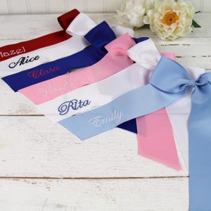 May include: A collection of personalized satin sashes in various colors, including red, blue, and pink. Each sash is embroidered with a different name in elegant script. The sashes are arranged with bows, suggesting a celebratory or event theme.