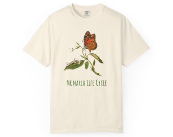 May include: A cream-colored t-shirt with a graphic of a monarch butterfly, caterpillar, and chrysalis on a green stem. The text "MONARCH LIFE CYCLE" is printed in green below the graphic.