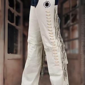 Peut inclure: Chaps en cuir blanc avec franges décoratives et détails en bois. Les chaps comportent une ceinture et un emblème décoratif sur le haut de la cuisse. Le modèle porte des bottes noires. Vêtements de style western.