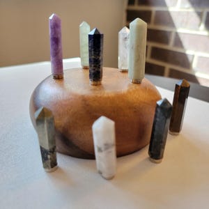 May include: A wooden bowl holds nine crystal points in various colors, including purple, green, and clear. The crystals have gold-colored metal bases. The bowl is set on a white surface, with a brick wall visible in the background.