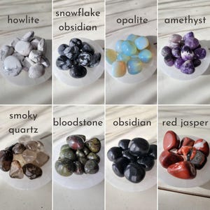 May include: Assortment of tumbled stones on white circular bases. Stones include howlite, snowflake obsidian, opalite, amethyst, smoky quartz, bloodstone, obsidian, and red jasper.