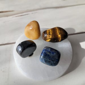 May include: Four tumbled stones on a white selenite charging plate. The stones are a yellow tiger's eye, a black obsidian, a blue lapis lazuli, and a brown tiger's eye.