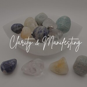 May include: A collection of polished gemstones in various colors, including blue, clear, yellow, and green, displayed in a white, shallow bowl. The text "Clarity & Manifesting" is written in cursive across the image. The stones are arranged on a white surface.