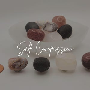 May include: A collection of smooth, polished stones in shades of black, white, pink, and mauve, arranged in a white bowl and on a surface next to a copper penny. The text "Self-Compassion" is overlaid in white script across the center of the image.