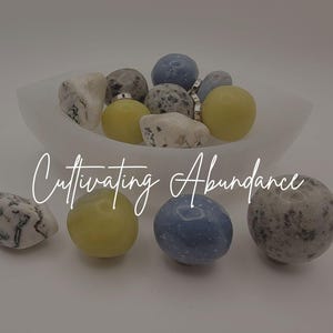 May include: A white translucent bowl filled with a collection of smooth, polished stones in shades of yellow, blue, and white with grey speckles. Several additional stones are arranged in front of the bowl. The text Cultivating Abundance is centered.