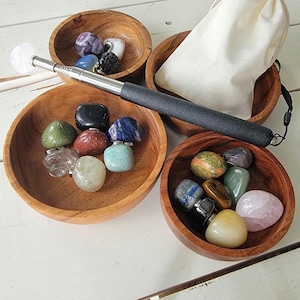 May include: A collection of polished gemstones in various colors, displayed in four wooden bowls. A white drawstring bag and a silver and black wand are also present. The gemstones include amethyst, quartz, and other colorful stones.