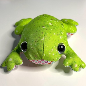 May include: A stuffed frog toy made of bright green fabric with a speckled pattern and large black plastic eyes. The underside of the frog features a white fabric with a colorful pattern of small pink and blue crosses and other shapes.