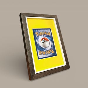 Puede incluir: Tarjeta Pokémon enmarcada con el diseño de la Poké Ball sobre fondo azul, enmarcada en un passe-partout amarillo. La tarjeta está en un marco de madera marrón oscuro con un borde interior blanco, para coleccionistas.
