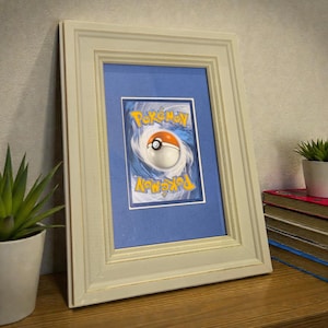 Puede incluir: Carta de Pokémon enmarcada con un passe-partout azul y un marco de madera color crema. La carta presenta un diseño de Poké Ball y la palabra "Pokémon". El marco está apoyado sobre una superficie de madera junto a una planta en maceta y una pila de libros.