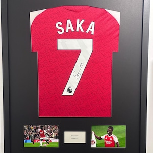 May include: Framed Arsenal football shirt, predominantly red with white detailing, displaying "SAKA" and the number "7". Includes a signature and two small photographs of the player. Presented in a black frame.
