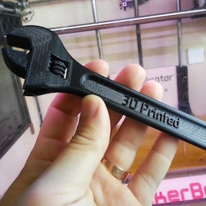 以下が含まれることがあります： 表面に質感があり、ハンドルに「3D Printed」の文字が浮き彫りになった黒いモンキーレンチ。可動式のジョー機構とローレット加工された調整ホイールを備えており、積層造形特有の層状の構造が見て取れるツールです。