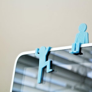 May include: Two light blue plastic figures designed to perch on the edge of a monitor. One figure is shaped like a person sitting with legs dangling, while the other figure is shaped like a person climbing up the side of the screen frame.