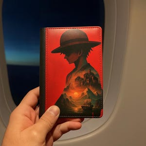 May include: Red and black passport holder featuring a silhouette of a character wearing a hat, with a landscape scene inside. The holder is held in front of an airplane window.