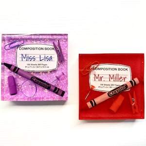 May include: Two resin paperweight desk organizers with a school theme. The left paperweight is purple and features a purple crayon, a purple pencil, a paperclip, and a push pin. The right paperweight is red and features a pink crayon, a red pencil, a paperclip, and a clothespin. Both paperweights have a white label that says "Composition Book" and a name. The left paperweight says "Miss Lisa" and the right paperweight says "Mr. Miller".