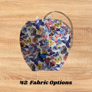 May include: A round, fabric-covered item with a butterfly pattern in shades of blue, pink, yellow, and purple. The item has a clear plastic handle and tubing. Text at the bottom reads "42 Fabric Options."