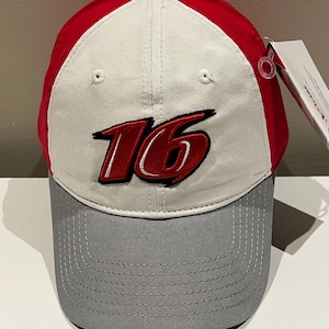 May include: A baseball cap featuring a red top, white central panel, and a grey brim. The number "16" is embroidered in red with a black outline. A tag is attached to the side.