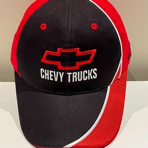 May include: A black and red baseball cap with the text "CHEVY TRUCKS" embroidered in white. The cap has a red brim and top, with a white stripe separating the red and black sections. The Chevrolet logo is displayed in red.