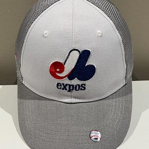 May include: A grey and white baseball cap with the Montreal Expos logo in red, white, and blue. The cap has a mesh back and a baseball pin on the side. The word "expos" is embroidered below the logo.