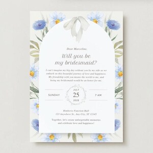 May include: A floral-themed bridesmaid proposal card with a white ribbon and blue flowers. The card reads "Dear Marceline, Will you be my bridesmaid?" and includes the date and time of the wedding.