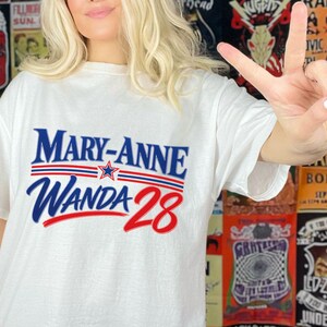 May include: A white short-sleeved t-shirt featuring the blue text MARY-ANNE above the red and blue text WANDA 28. The design includes a small blue star centred between two horizontal red and blue lines, creating a bold graphic print.
