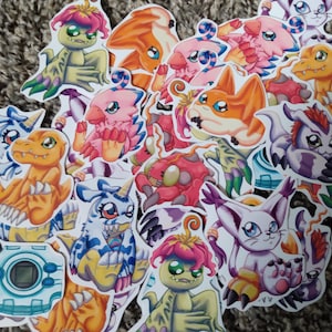 May include: A collection of colorful cartoon character stickers. The stickers feature various creatures in shades of orange, pink, blue, and green. Some stickers have a digital camera design.