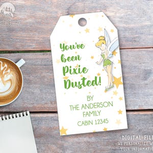 May include: A white printable gift tag with a green and yellow illustration of Tinkerbell. The tag says "You've been Pixie Dusted!" with the text "By The Anderson Family Cabin 12345" below the illustration.