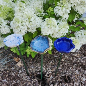 May include: Three ceramic garden art pieces in the shape of flowers. One is white with blue speckles, one is light blue, and the third is a deep blue. Each flower is mounted on a green metal stake, perfect for adding a touch of color to any garden.