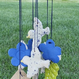 May include: A ceramic wind chime with blue, white, and green painted ceramic pieces. The pieces include a bird, a flower, and a leaf. The wind chime is hanging from a string.