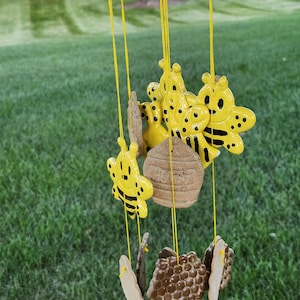 May include: A yellow and brown ceramic wind chime featuring bees and honeycombs. The chime hangs from a wooden branch.