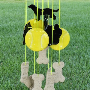May include: A ceramic wind chime featuring a black dog silhouette, yellow tennis balls, and beige bone shapes. The wind chime is hanging from a string and is in front of a green grassy background.