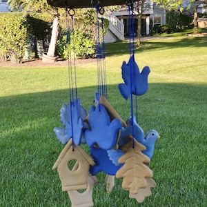 May include: A wooden wind chime with blue ceramic birds and a birdhouse, a leaf, and a tree hanging from it. The wind chime is hanging from a tree branch in a backyard.