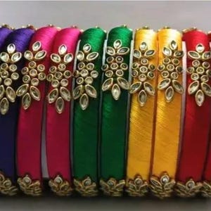 May include: A collection of colorful bangles in shades of purple, pink, green, yellow, and red. Each bangle is adorned with gold-colored embellishments and clear gemstones, creating a decorative and eye-catching design.