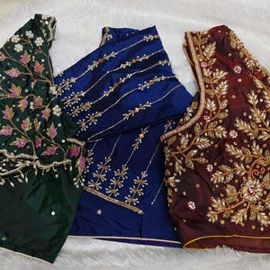 May include: Three intricately embroidered blouses in jewel tones. The first is dark green with floral and leaf patterns in white, pink, and gold. The second is a deep blue with gold linear and starburst designs. The third is burgundy with gold floral embroidery.