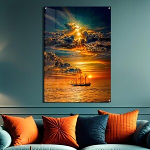 Pode incluir: Uma peça de arte de parede retangular apresentando um veleiro no oceano ao pôr do sol. O céu está repleto de nuvens dramáticas iluminadas pela luz dourada do sol, com raios descendo. A água reflete os tons alaranjados e amarelos do sol.